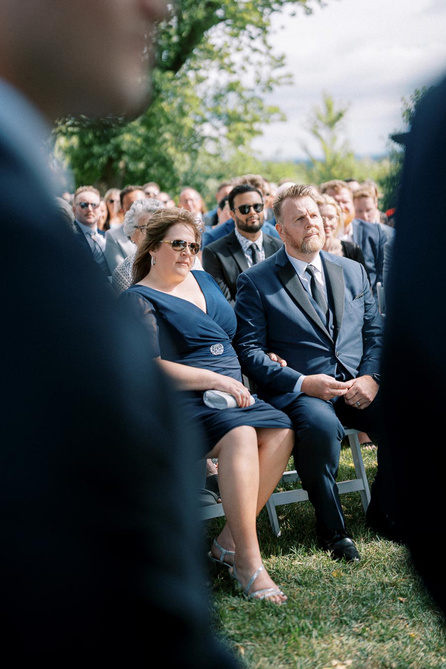 Guests seated outdoors at a wedding ceremony, attentively watching the event on a sunny day, with trees in the background and everyone dressed in formal attire.