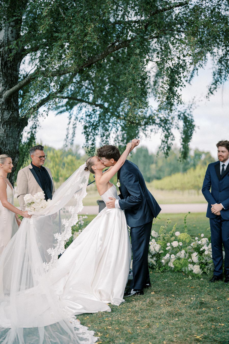 A bride and groom share a kiss under a tree during a beautiful outdoor wedding ceremony, with bridesmaids and groomsmen standing nearby, surrounded by lush greenery and floral arrangements.