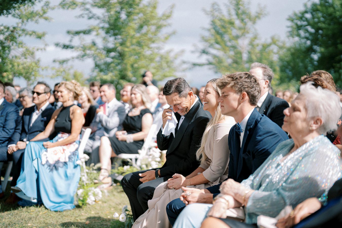 Guests sitting at an outdoor wedding ceremony, with one man wiping a tear, dressed in formal attire, surrounded by lush greenery on a sunny day.
