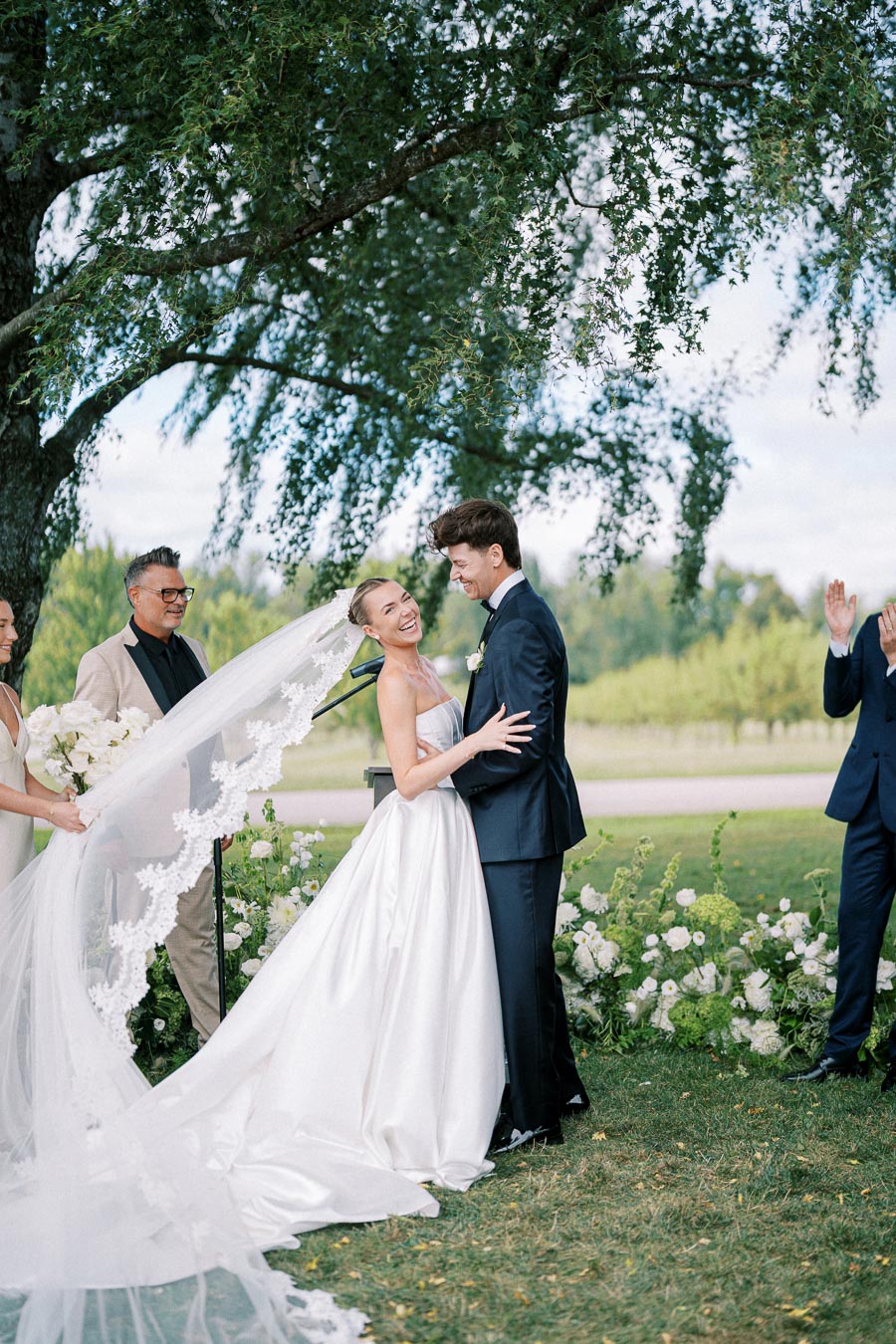 A joyful bride and groom stand together under a lush tree during their outdoor wedding ceremony, surrounded by greenery and floral arrangements. The bride wears a flowing white gown with a long veil, while the groom is dressed in a dark suit. A wedding officiant is present in the background, contributing to the celebratory atmosphere.