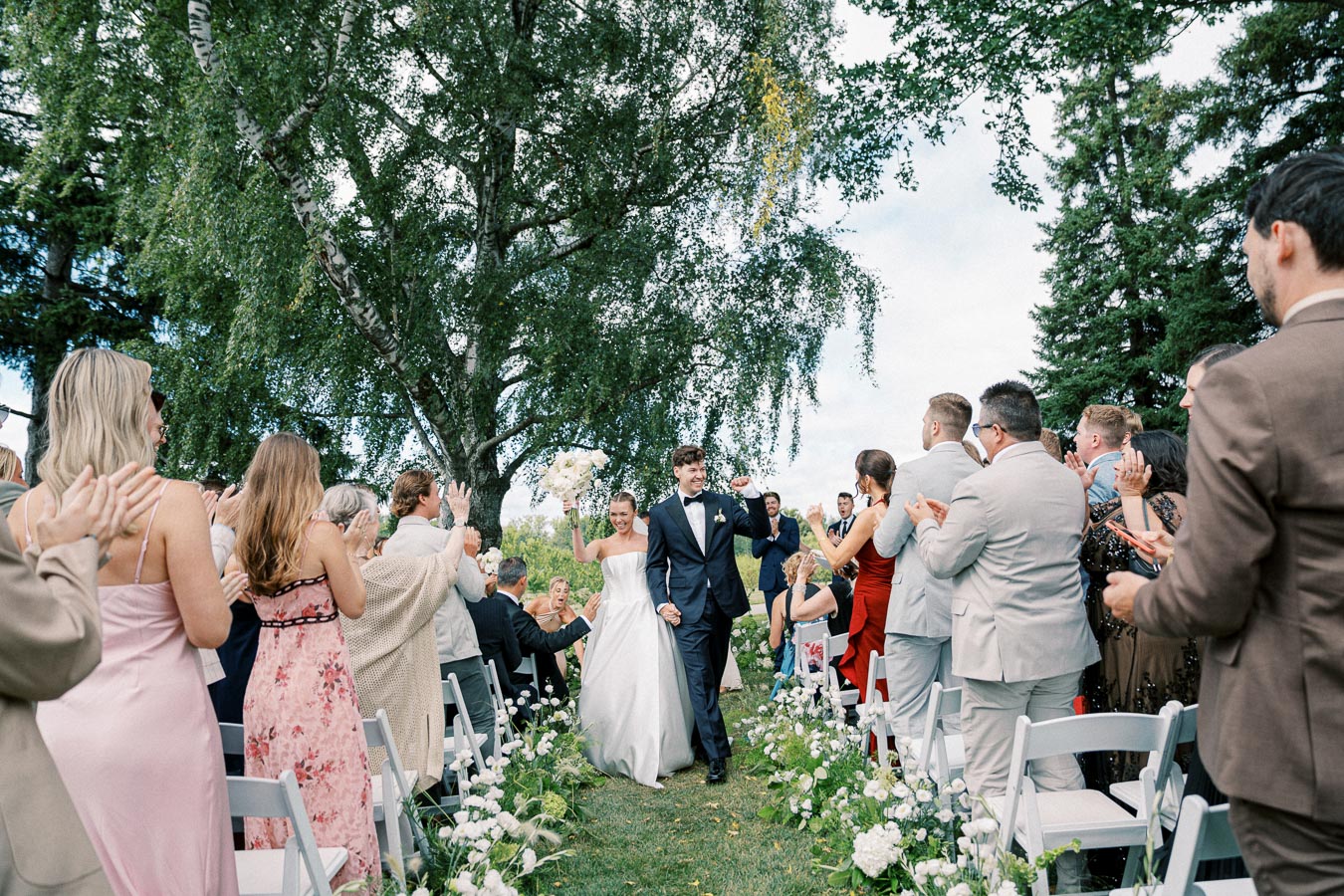 Outdoor wedding ceremony with newlywed couple walking down the aisle, surrounded by cheering guests on a sunny day, with lush greenery and floral decorations.