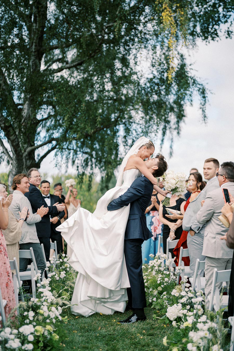 A bride and groom share a joyful embrace during their outdoor wedding ceremony, surrounded by applauding guests and lush greenery.