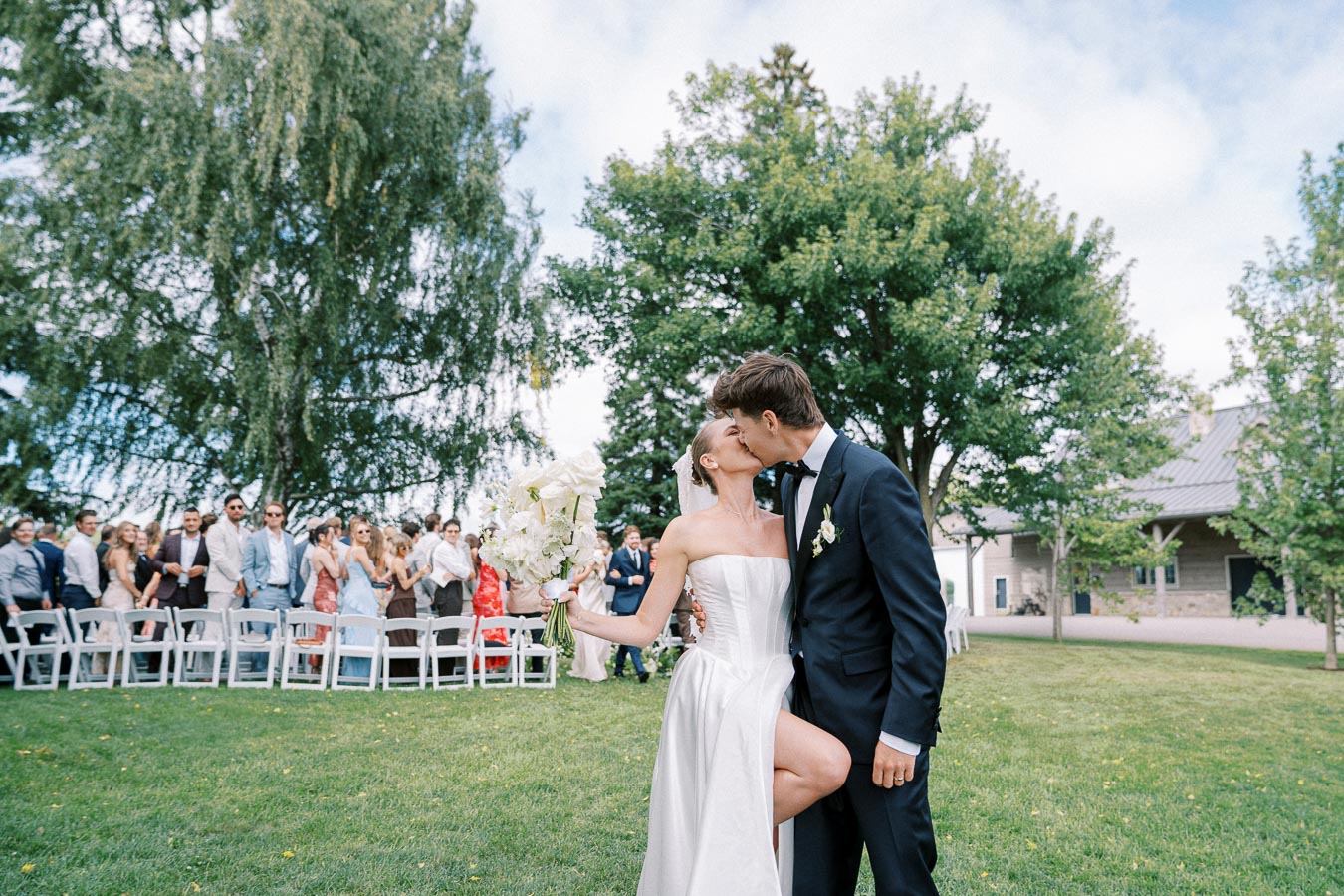 Bride and groom share a kiss outdoors during a wedding ceremony, surrounded by guests under a clear sky with lush greenery.