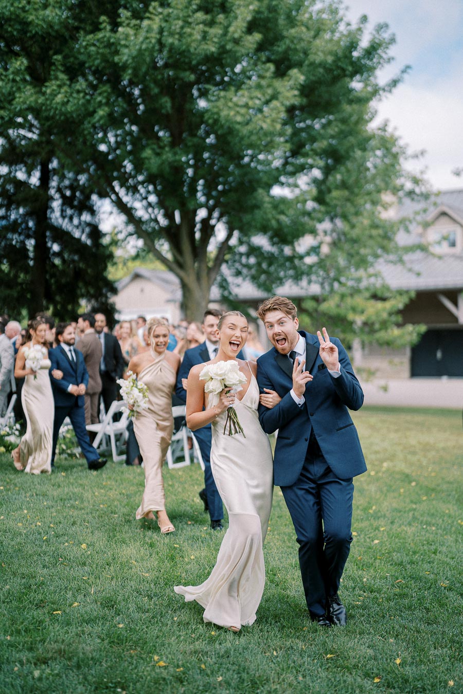 A joyful wedding party exits the ceremony outdoors, with bridesmaids in champagne dresses holding bouquets, and groomsmen in navy suits celebrating on a lush green lawn under a clear sky.