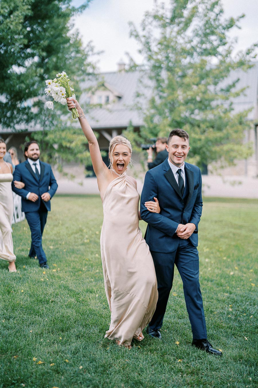 A joyful bridesmaid in a cream dress holding a bouquet walks arm-in-arm with a smiling groomsman in a navy suit on a lush green lawn. Other members of the wedding party are seen in the background, celebrating outdoors on a sunny day.