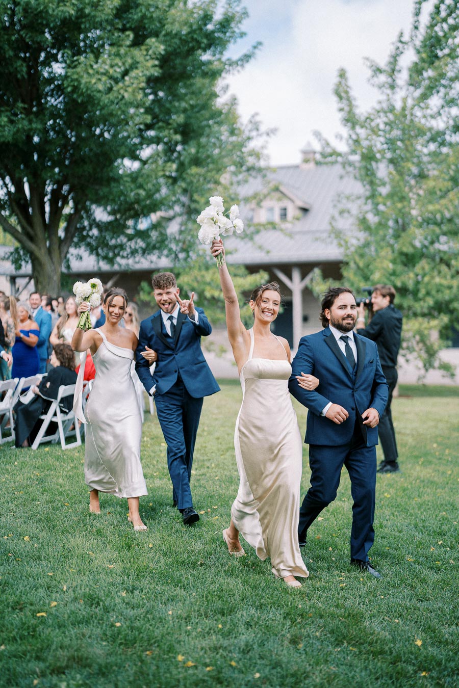 A joyful outdoor wedding procession with elegantly dressed bridesmaids in white dresses and groomsmen in navy suits, holding flowers and celebrating in a lush garden setting.