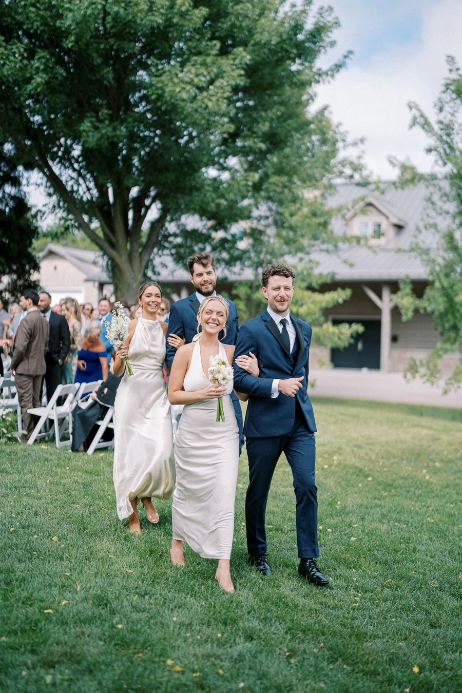 Wedding party walking down the aisle during an outdoor ceremony, with bridesmaids in ivory dresses and groomsmen in navy suits, surrounded by lush greenery and guests seated on white chairs.