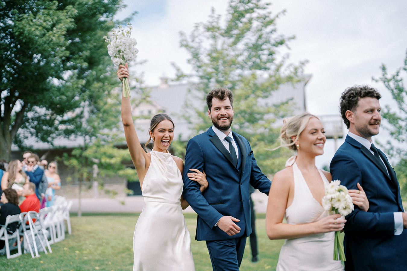 A group of happy wedding guests in formal attire walking outdoors, with a woman joyfully holding a bouquet of flowers and others smiling, surrounded by greenery and seating.