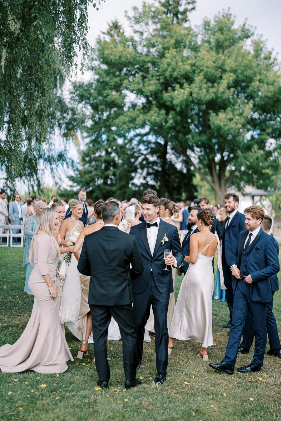 A group of elegantly dressed wedding guests socialize outdoors on a sunny day, surrounded by lush green trees and wedding decor. The men wear suits and bow ties, while the women are in flowing dresses, creating a sophisticated and celebratory atmosphere.