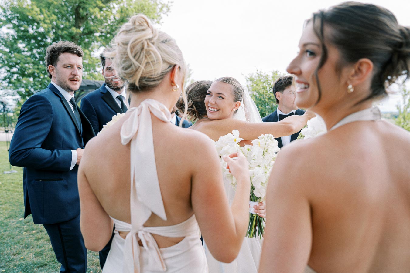 Wedding celebration with guests dressed in formal attire; a bride smiling and hugging a friend outdoors, surrounded by bridesmaids in cream dresses and groomsmen in suits, with greenery in the background.