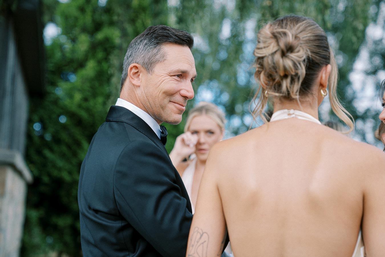 A man smiling in a tuxedo interacts with a woman in a white halter dress at an outdoor event, surrounded by greenery.
