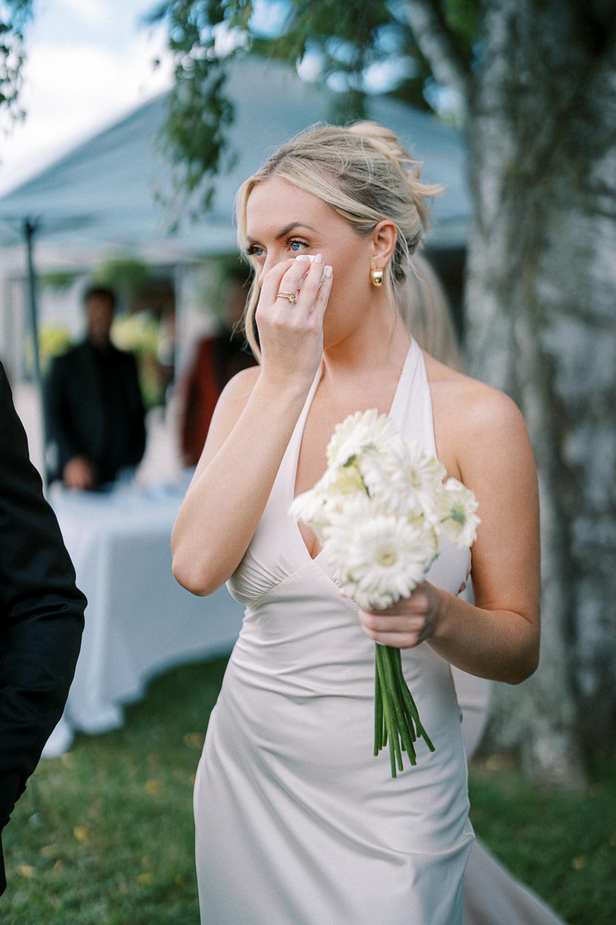 A bride in an elegant white dress holding a bouquet of white flowers, standing outdoors with a tearful, emotional expression during a wedding ceremony.