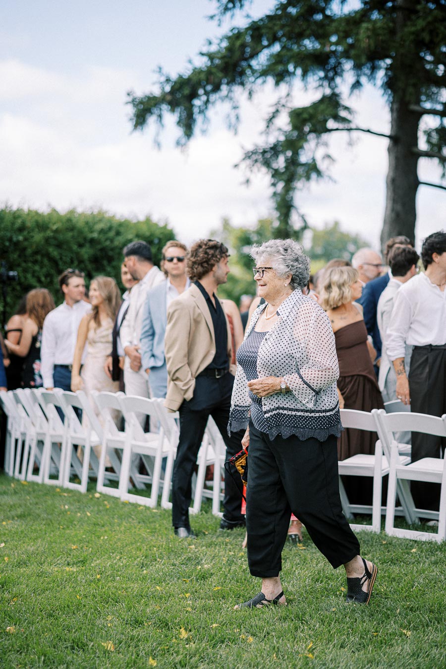 Elderly woman walking past seated guests at an outdoor wedding ceremony on a sunny day, with attendees dressed in formal attire and trees in the background.