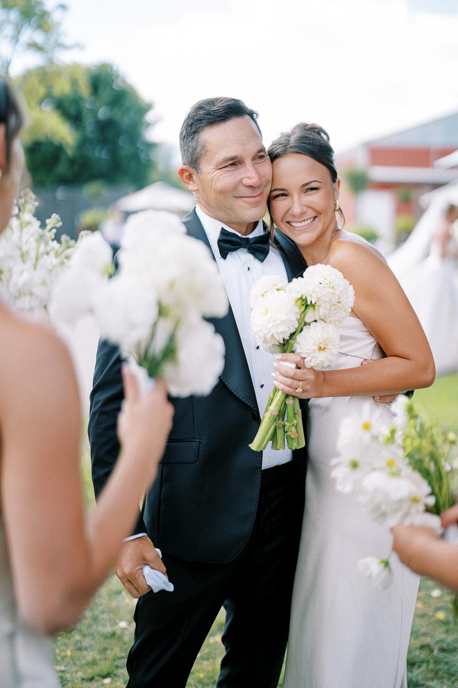 A joyful bride in a white dress embraces a man in a tuxedo, both holding white flower bouquets, surrounded by bridesmaids, during an outdoor wedding celebration.