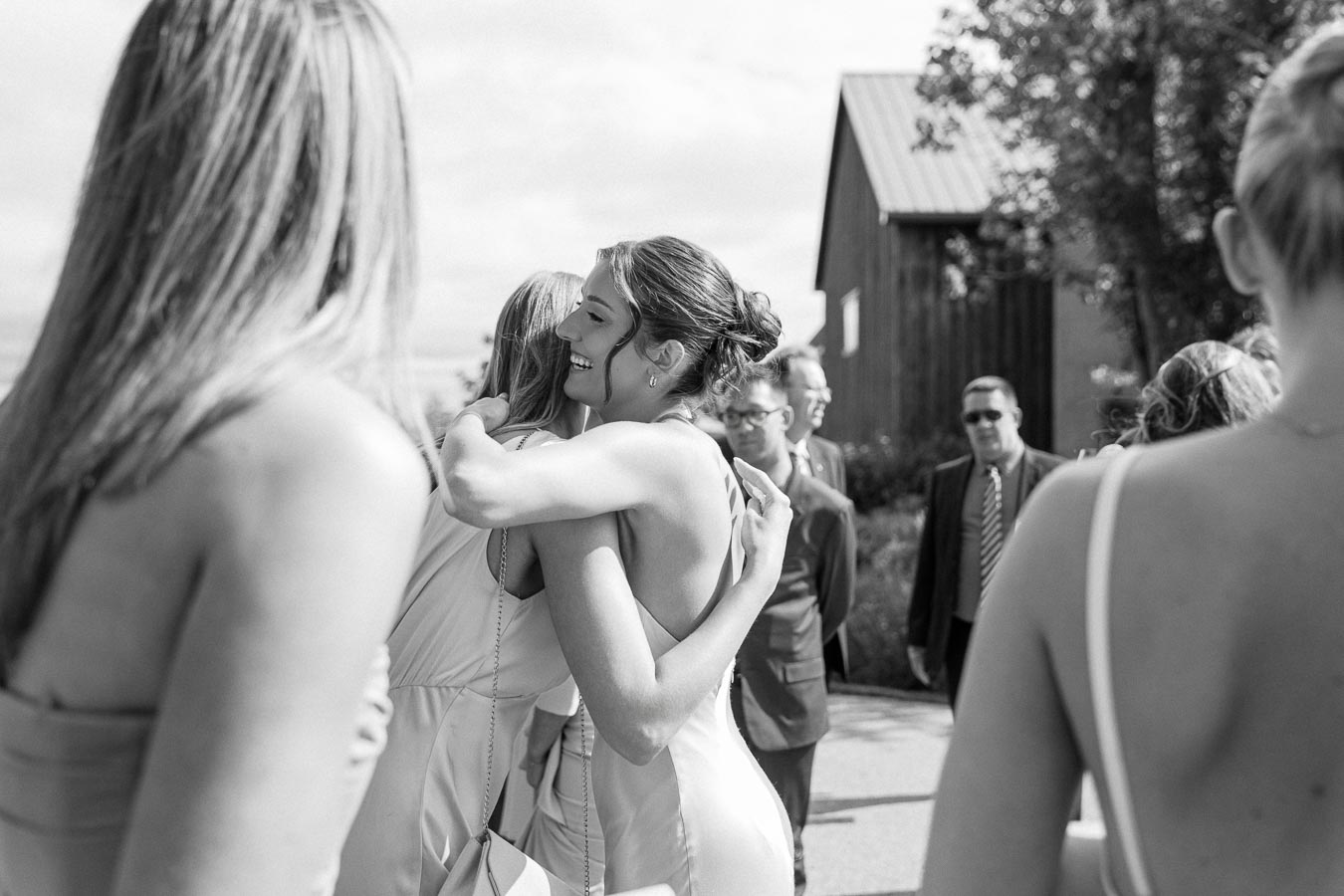 Black and white photo of a woman in a sleeveless dress warmly hugging another person outdoors at a wedding ceremony, with people in formal attire and a rustic wooden building in the background.