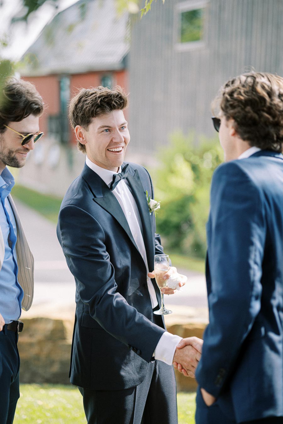 A groom in a formal blue suit shaking hands with a guest at an outdoor wedding reception, holding a glass of champagne, with a rustic barn in the background.