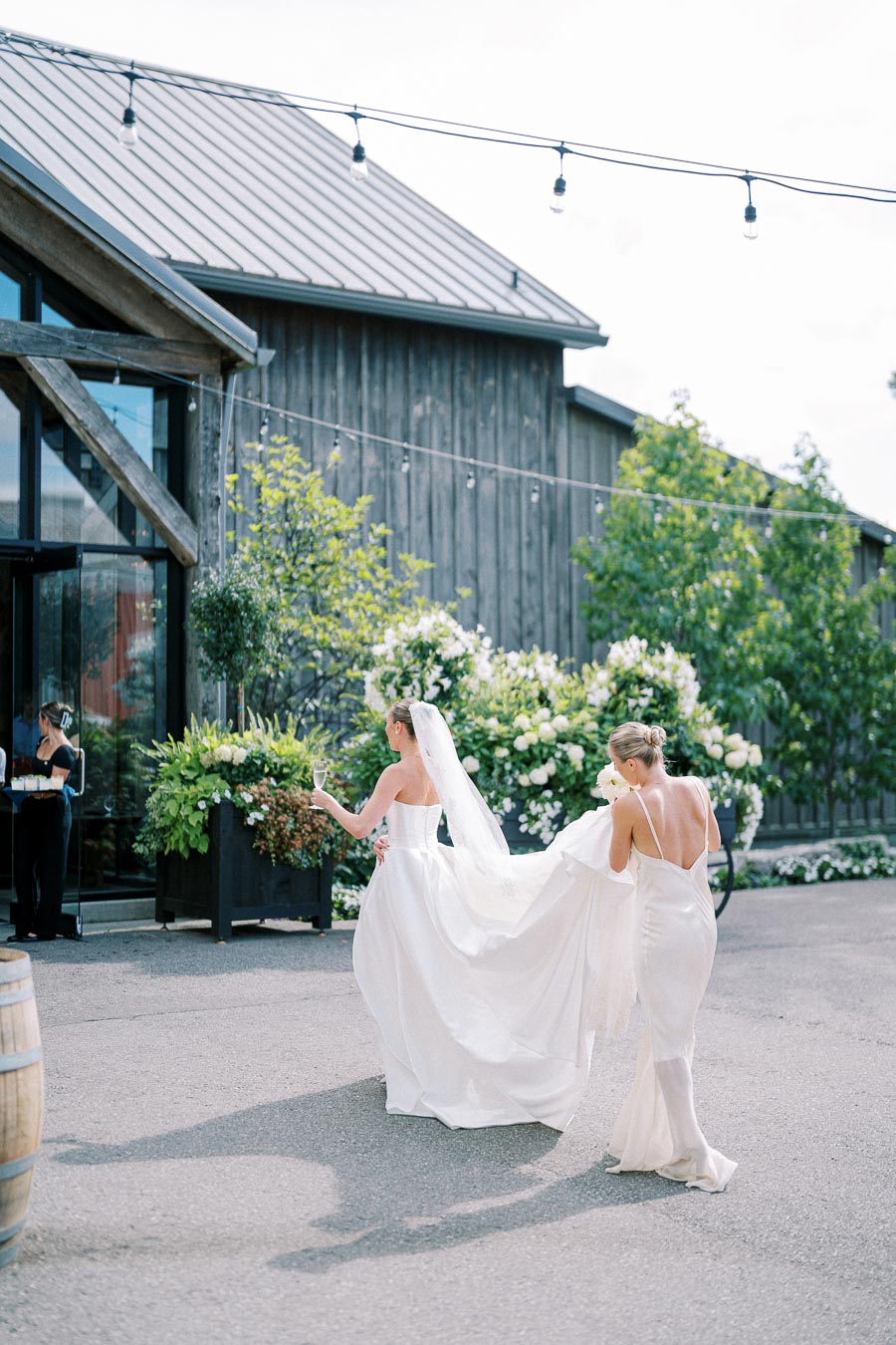 Wedding scene with bride in flowing white gown and veil, walking outside rustic venue, holding a glass of champagne, assisted by a woman in a satin dress, surrounded by greenery and white flowers under string lights.