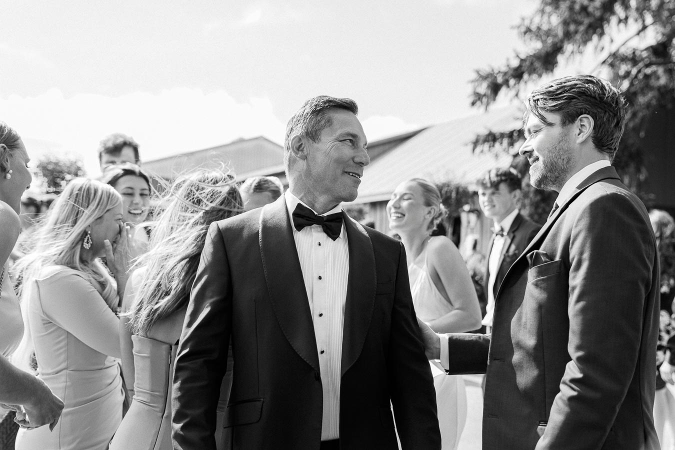 Black and white photo of a group of people dressed in formal attire, laughing and socializing at an outdoor event, possibly a wedding or celebration. The central figure is a man in a tuxedo, engaging in conversation and surrounded by smiling guests.