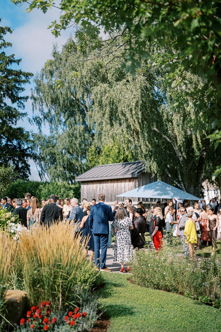 A group of people at an outdoor garden party, dressed in formal attire, under a clear blue sky with lush green trees and vibrant flowers in the background.