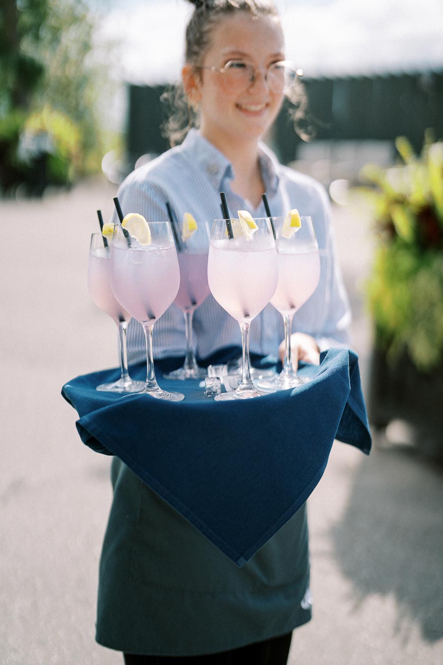A smiling server carrying a tray of refreshing pink cocktails garnished with lemon wedges and black straws, set on a sunny outdoor terrace.