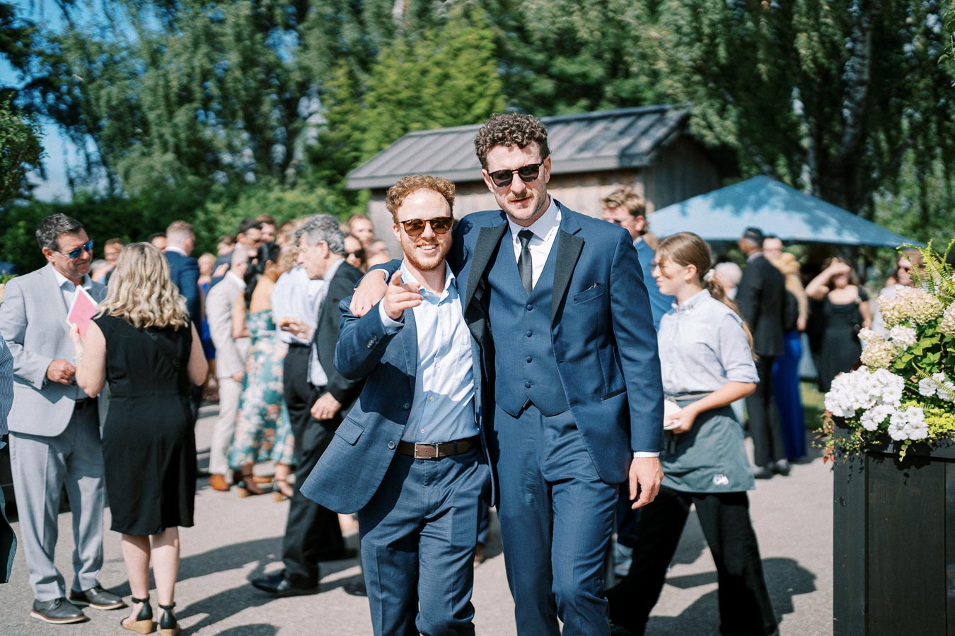 Two men in blue suits smiling and posing together at an outdoor event, surrounded by a crowd of people dressed in formal and casual attire with greenery and a building in the background.