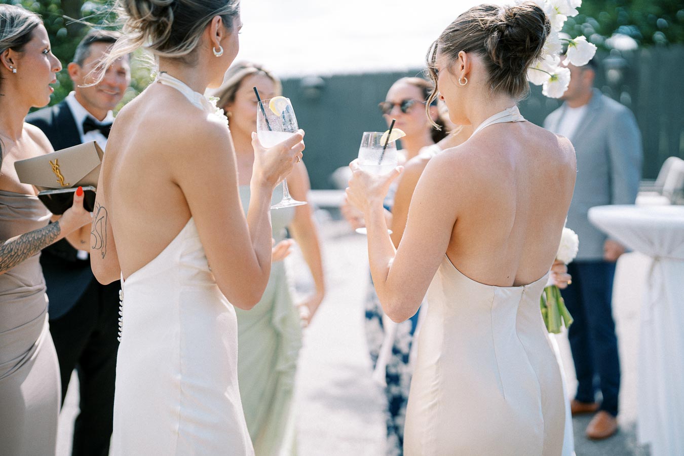 Elegant wedding guests in stylish dresses enjoying cocktails outdoors during a sunny celebration.