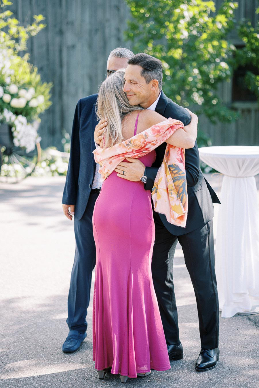 A man in a black suit embraces a woman in a vibrant pink dress outdoors, surrounded by greenery and sunlight, conveying warmth and joy.