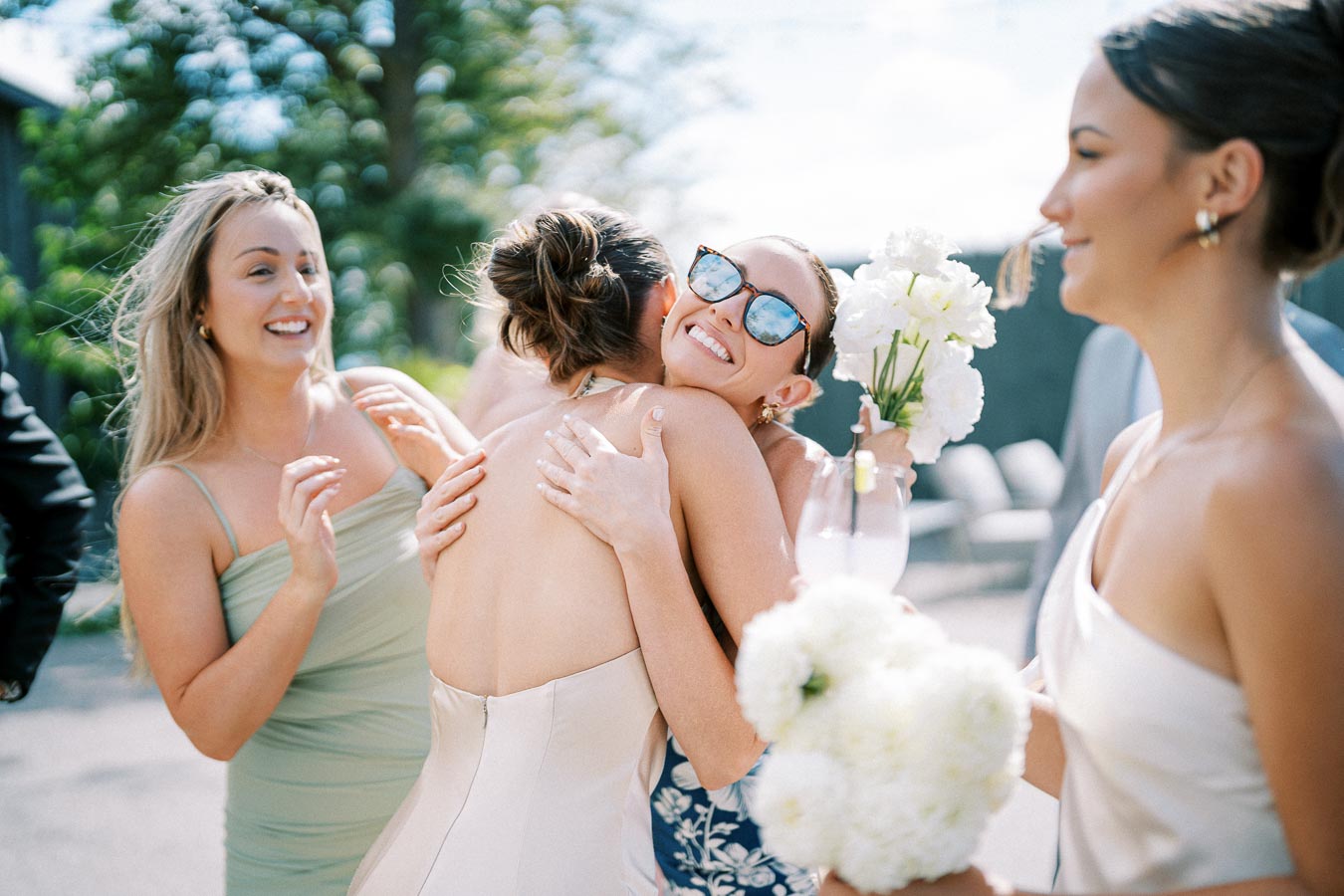 A joyful wedding scene with a bride embracing a guest holding flowers, surrounded by smiling bridesmaids in elegant dresses under a sunny sky.