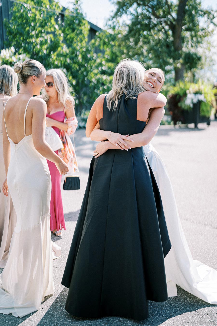 A joyful bride embraces a guest in a black dress at an outdoor wedding celebration, surrounded by elegantly dressed women under sunny skies and lush greenery.