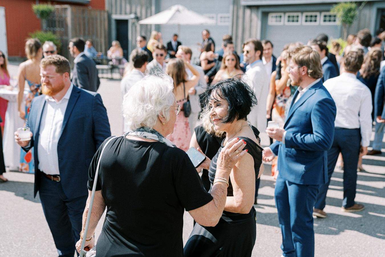 A diverse group of people socializing at an outdoor event, with individuals dressed in formal attire, engaging in conversation and enjoying drinks in a sunlit setting.