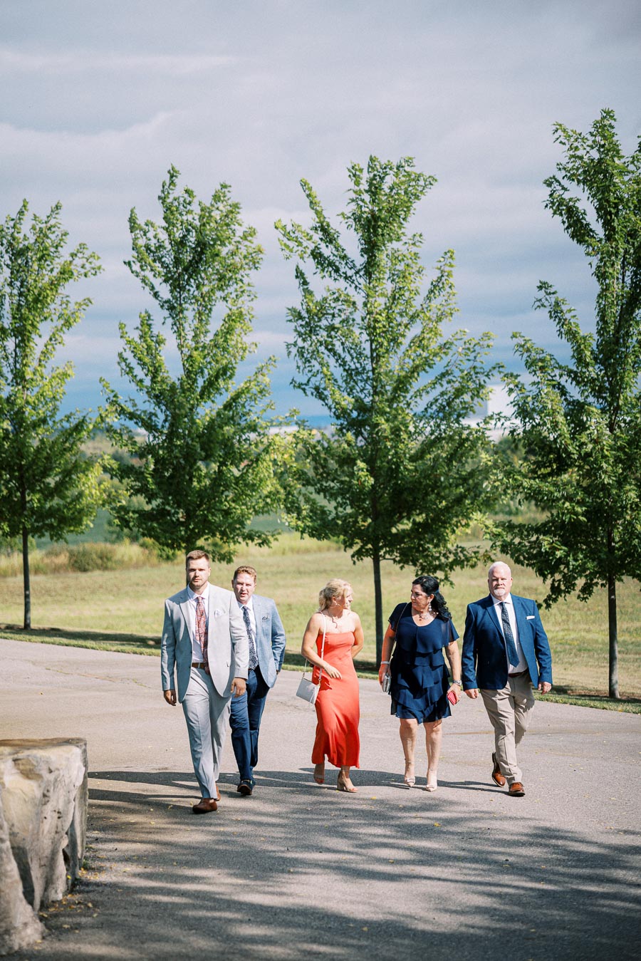 Group of people dressed in formal attire walking on a tree-lined pathway under a cloudy sky.