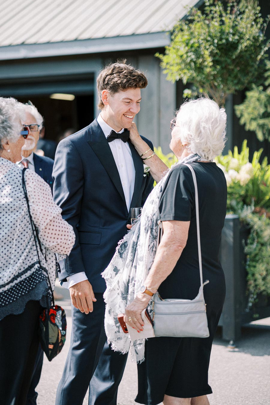A smiling man in a tuxedo interacts warmly with three older adults, capturing a joyful outdoor gathering.