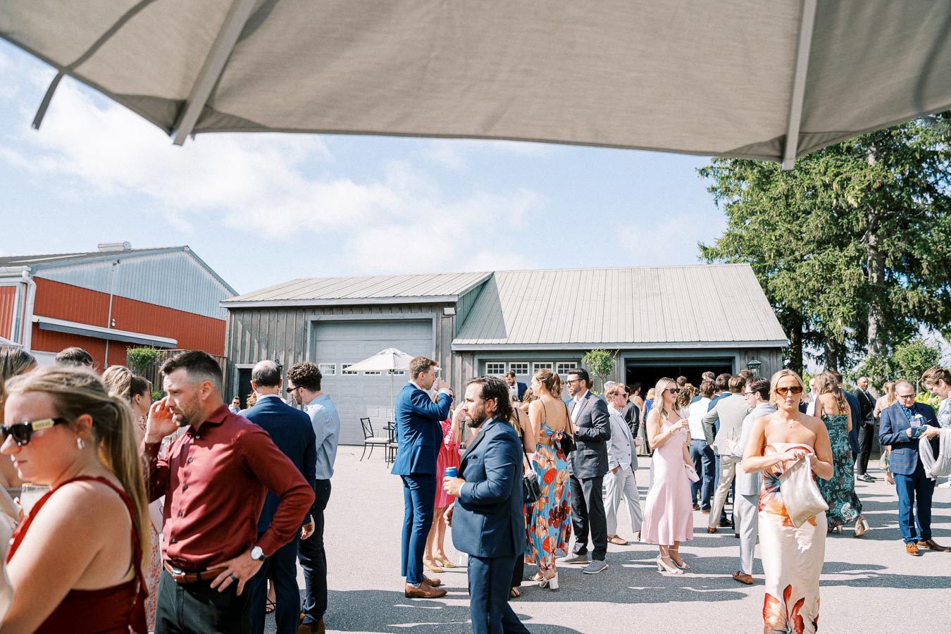 Outdoor social gathering with people dressed in formal and summer attire, mingling under a sunny sky near a rustic barn.
