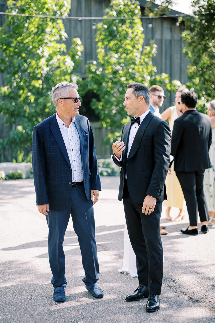Two men in formal attire engage in conversation outdoors, with a scenic backdrop of greenery. One wears sunglasses and a patterned shirt, and the other is in a black suit and bow tie. A group of people is visible in the background.