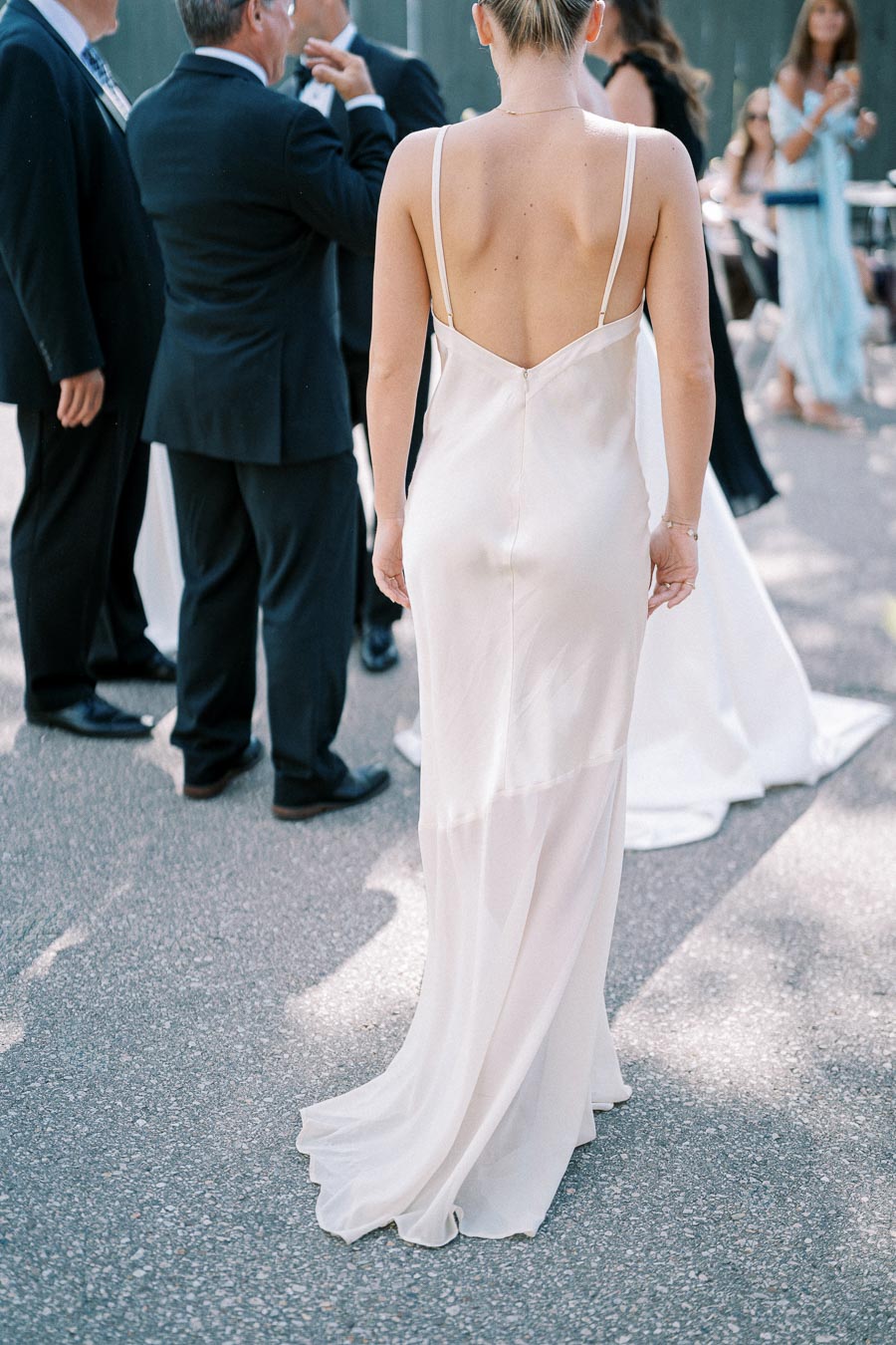 A woman in a backless ivory gown stands outdoors, surrounded by people in formal attire during a sunny event.