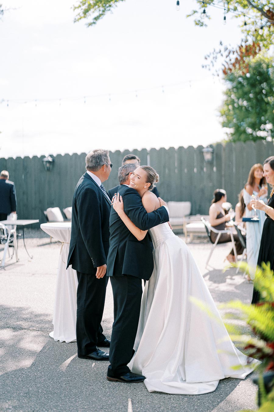 A bride in a white wedding dress warmly embraces a guest in a suit at an outdoor wedding reception, with other guests enjoying the celebration in the background.