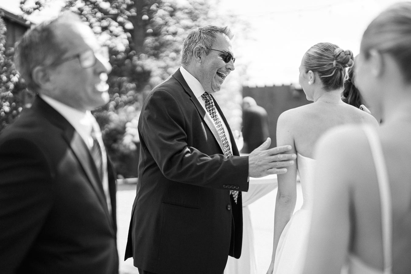 Black and white photo of a joyful wedding scene with a man in a suit laughing and engaging with a bride in a strapless dress, surrounded by other elegantly dressed guests outdoors.
