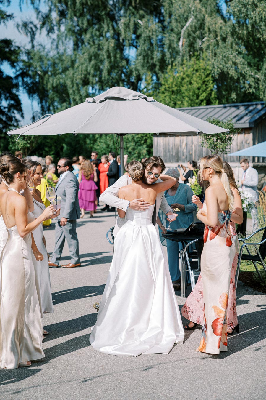 Outdoor wedding reception with guests mingling under umbrellas. A person in a white dress is hugging someone in sunglasses while other attendees in formal attire enjoy drinks and conversations. Trees and rustic buildings create a scenic backdrop.