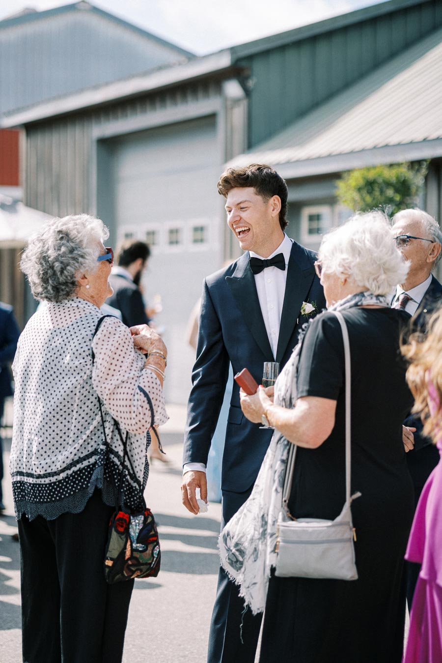 A young man in a tuxedo smiling and chatting with guests at an outdoor gathering, in front of a rustic building, celebrating a special occasion.
