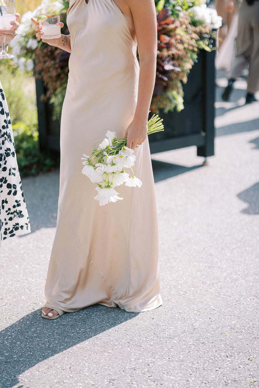 Elegant woman in a beige silk dress holding a bouquet of white flowers during an outdoor event.