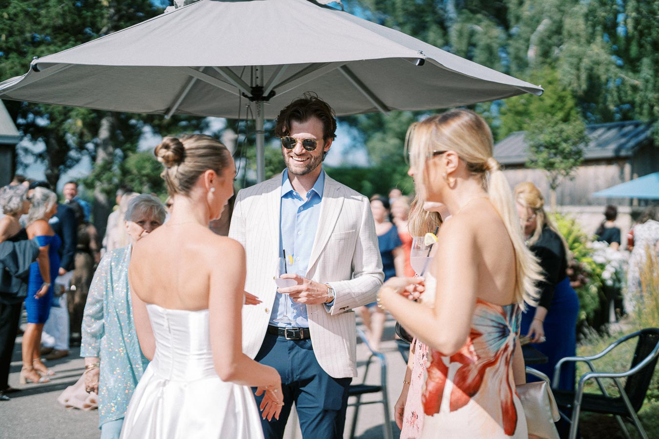 Group of elegantly dressed people enjoying a sunny outdoor event under a large patio umbrella, engaged in conversation with drinks in hand.