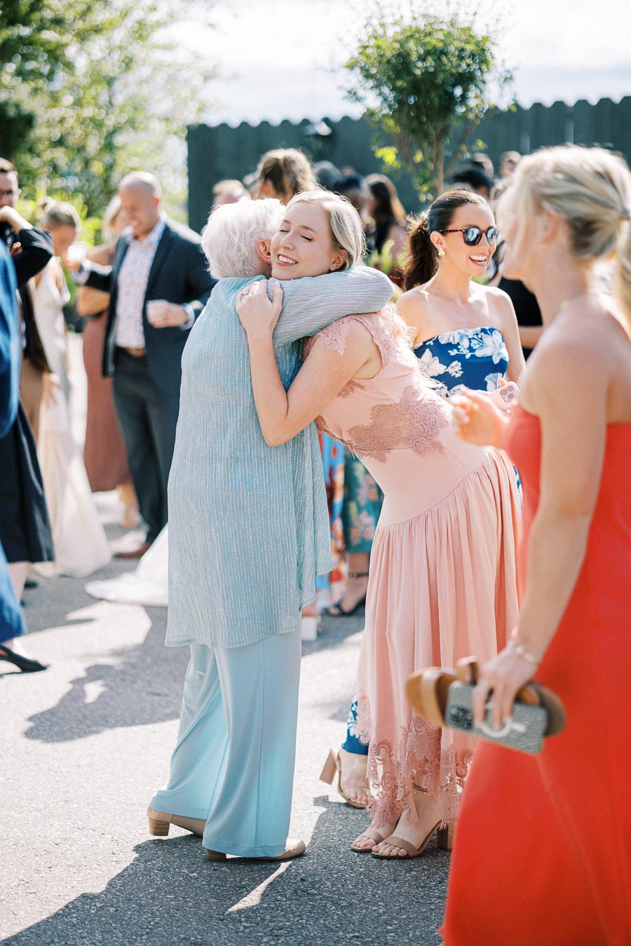 Guests embracing at an outdoor wedding ceremony, featuring a woman in a pink dress and another in a light blue outfit surrounded by smiling attendees in summery attire.