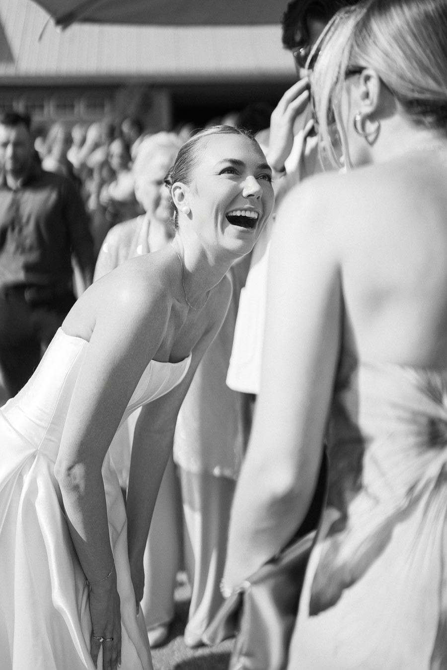 Black and white photo of a bride laughing joyfully at an outdoor wedding reception, surrounded by guests in elegant attire.
