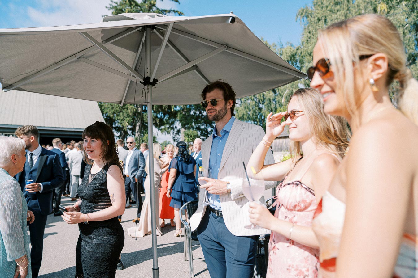 A group of elegantly dressed people enjoying a sunny outdoor social event, gathered under a large sunshade, with trees and a building in the background.