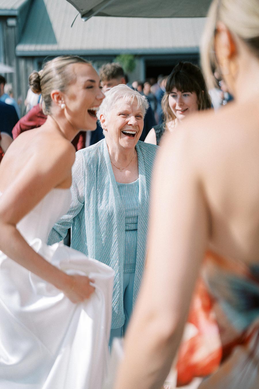 Group of women joyfully laughing at an outdoor wedding reception, with the bride wearing a white dress and an elderly woman in a light blue outfit.
