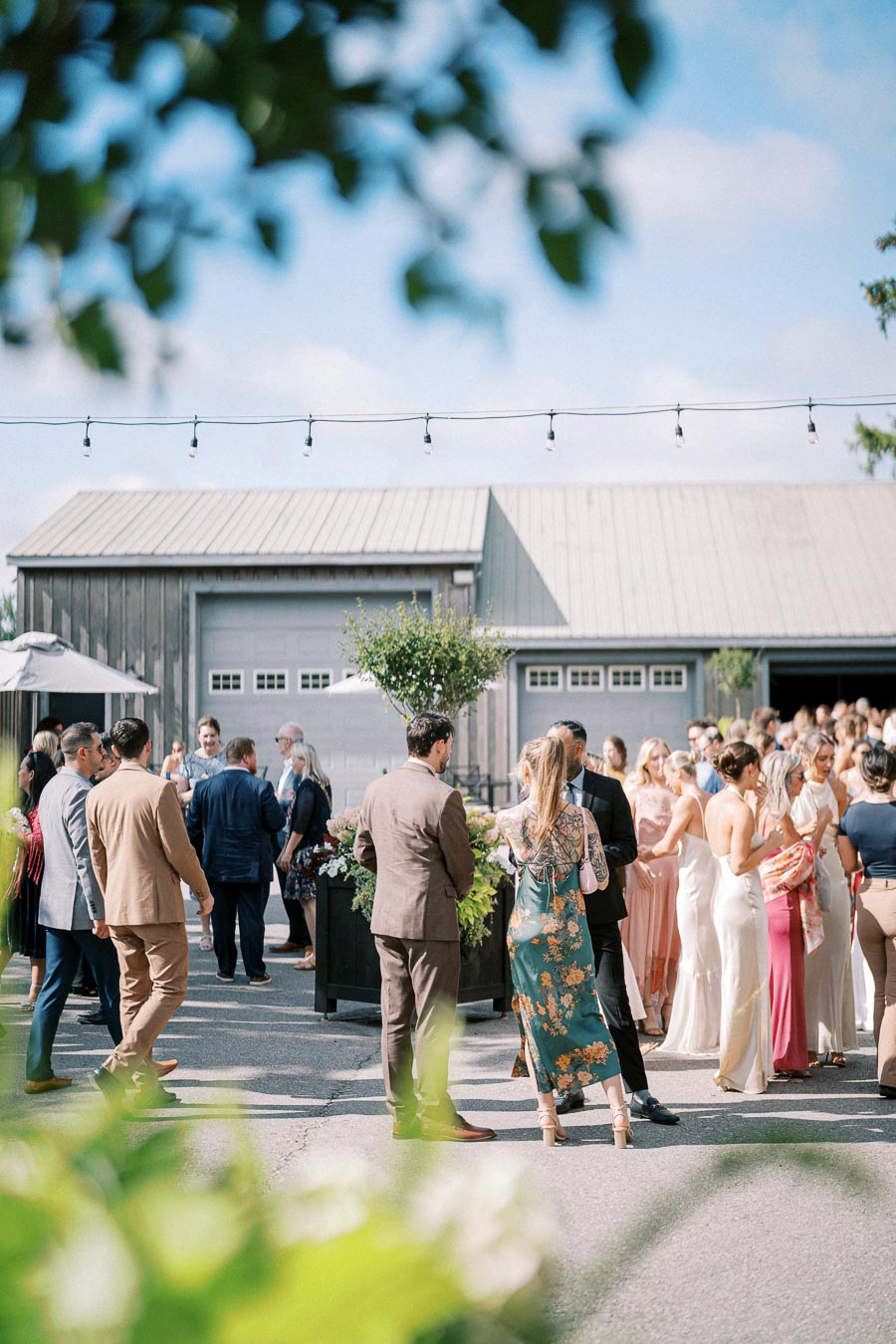 Outdoor wedding reception at a rustic venue, with well-dressed guests socializing under a clear blue sky.
