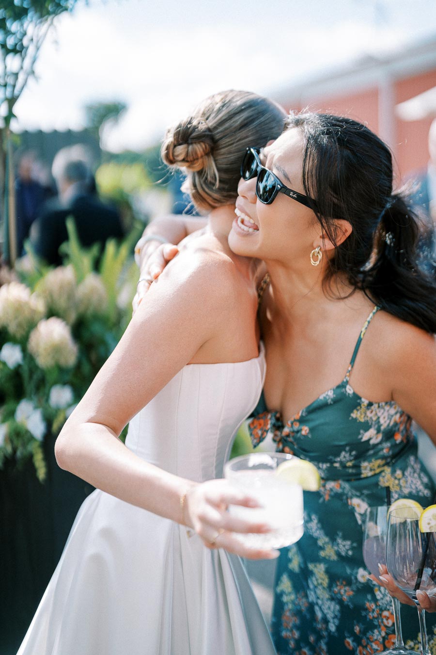 Two women embracing at an outdoor wedding celebration, with one wearing sunglasses and a floral dress, smiling warmly. The other woman is in a white dress with her hair in an elegant updo. The setting has a festive atmosphere with people and greenery in the background.