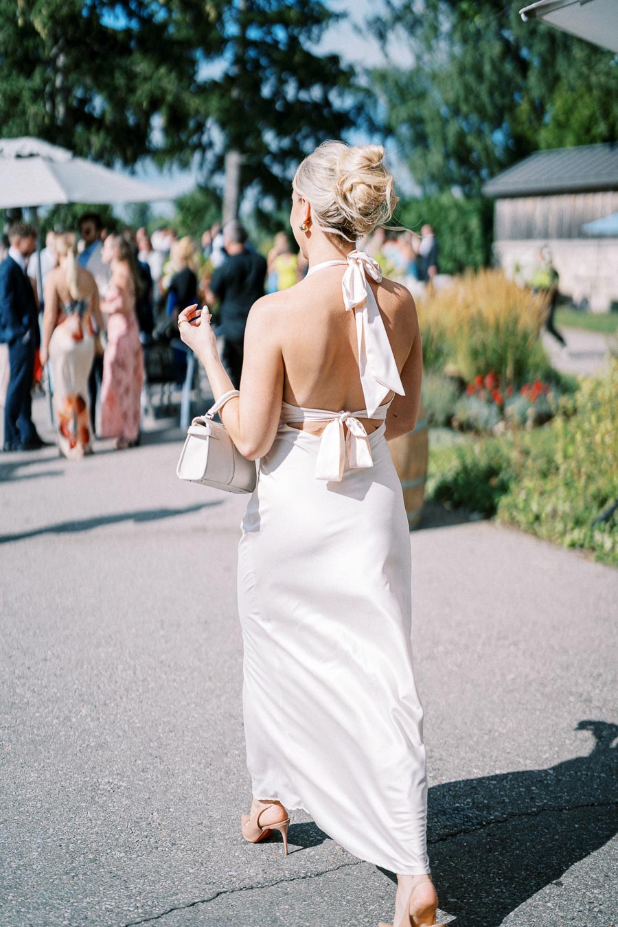 Elegant woman in a backless white dress attending an outdoor event, carrying a stylish cream handbag with people and greenery in the background.