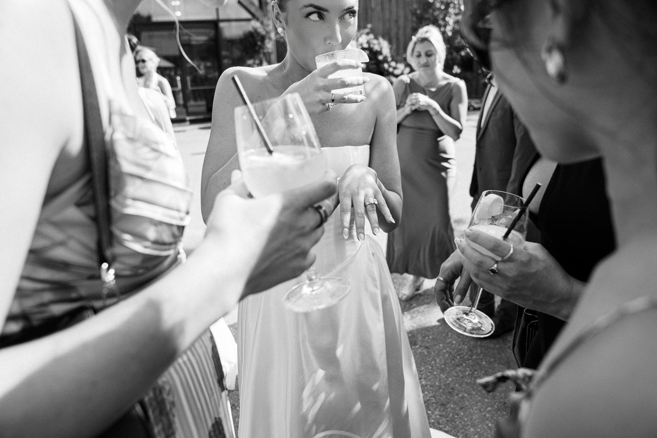 A group of women at an outdoor event enjoying drinks, with one woman in a dress showing off her rings and sipping from a glass, captured in black and white for a candid moment.