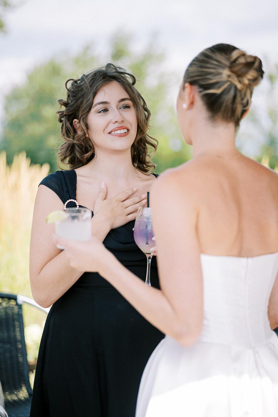 Two women at an outdoor event, one in a black dress and the other in a white dress, having a conversation while holding cocktails, with a blurred natural background.